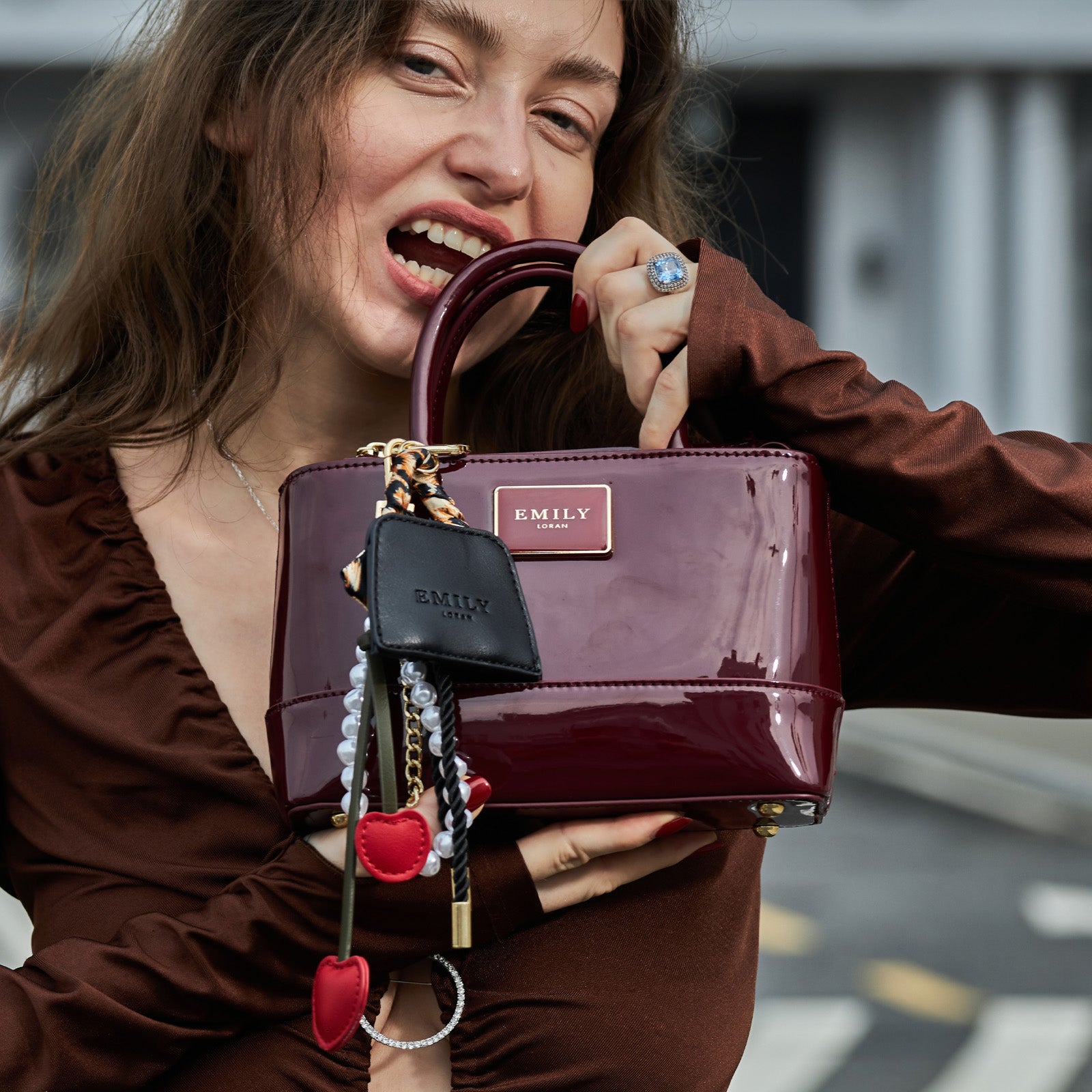 Woman in a brown top holding a glossy burgundy Emily Loran handbag with decorative keychains, smiling confidently.